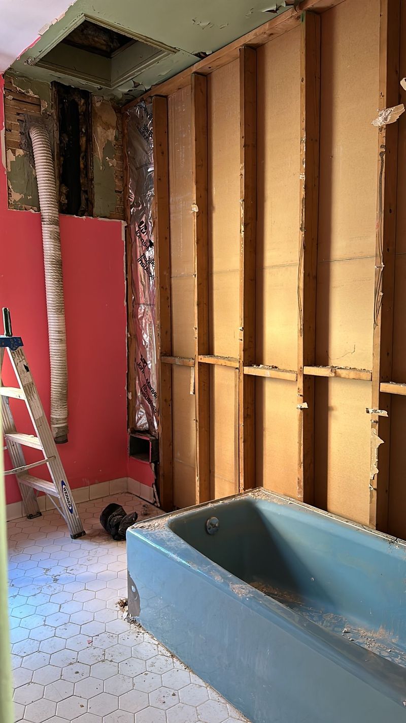 The original bathroom has a blue built in tub, pink walls, and exposed wall framing.